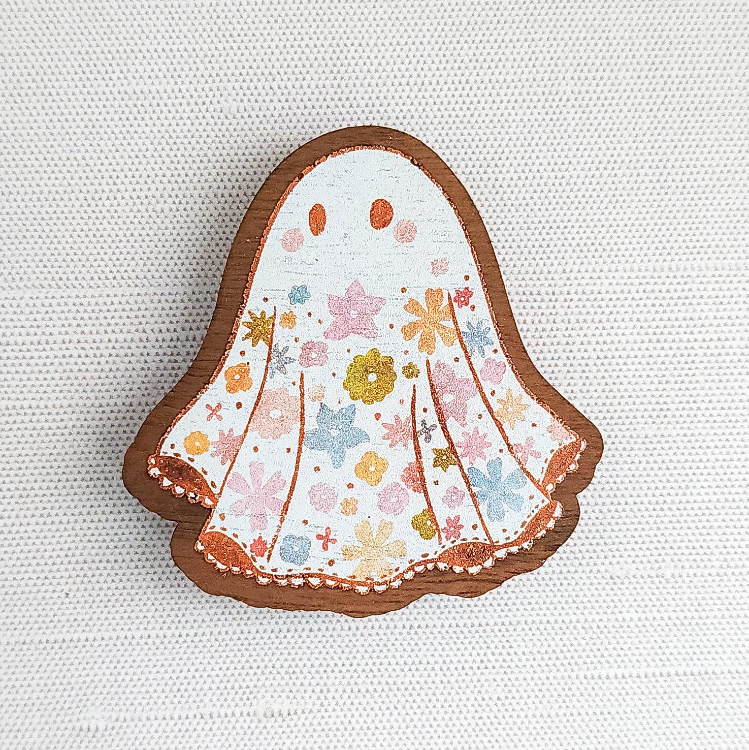 Wooden magnet featuring a little floral ghost with colorful flowers and stars on its sheet, printed on walnut wood and photographed on a neutral fabric background.