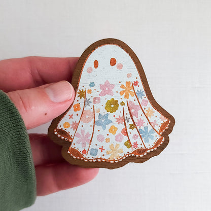 Wooden magnet featuring a little floral ghost with colorful flowers and stars on its sheet, printed on walnut wood and photographed held in a hand on a neutral fabric background.