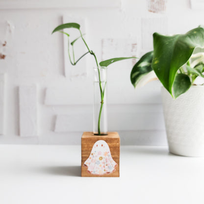 wooden block propagation station printed with a floral ghost. It has a glass tube with a green plant cutting on a white background.