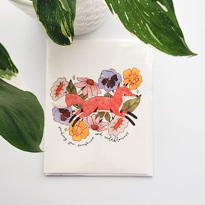 greeting card with an illustrated fox jumping through blooming flowers and the words sending you sunshine and wildflowers. The card is on a white background with a potted plant behind it