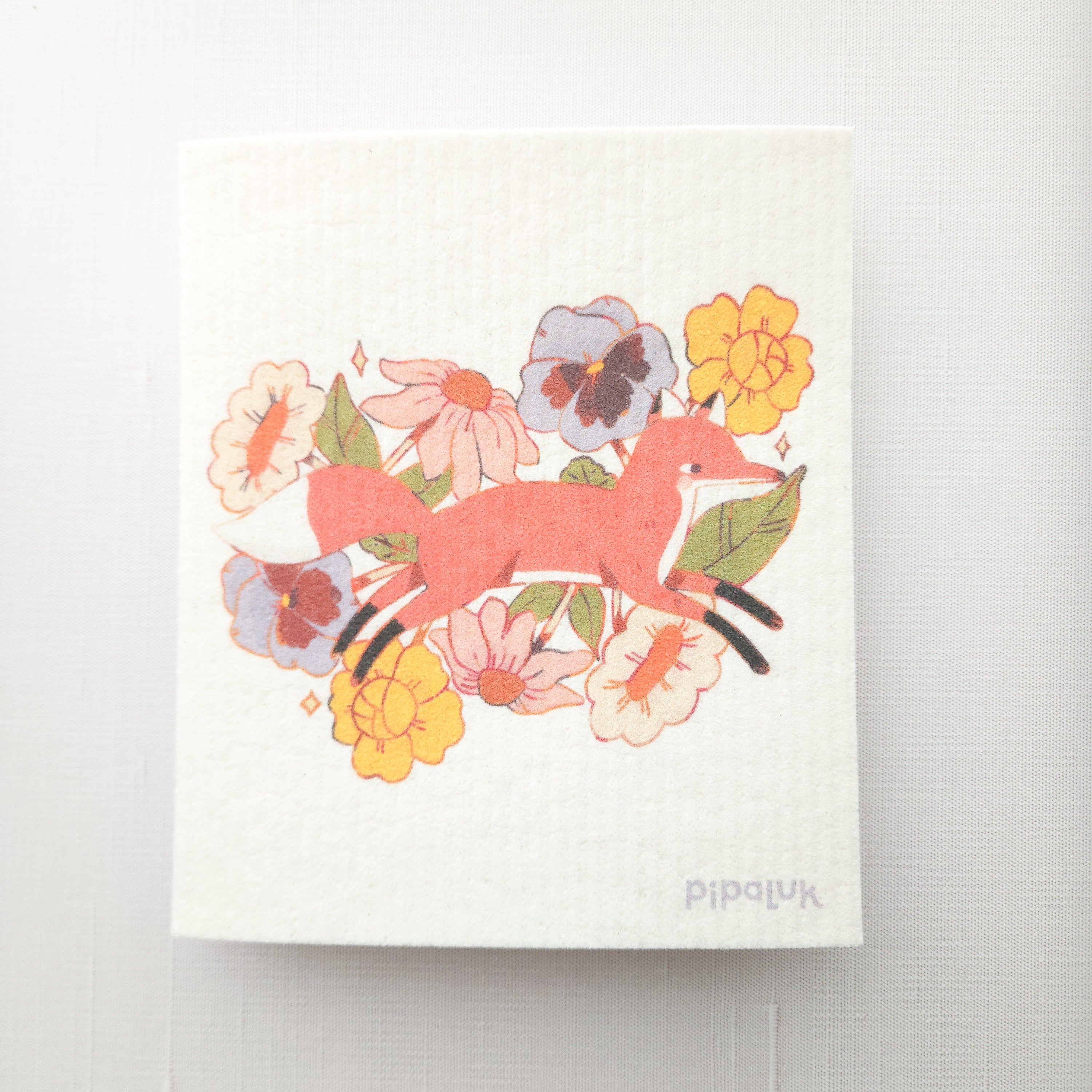 swedish dishcloth with an illustration of a fox jumping through flowers and a pipaluk logo in the bottom corner on a white background