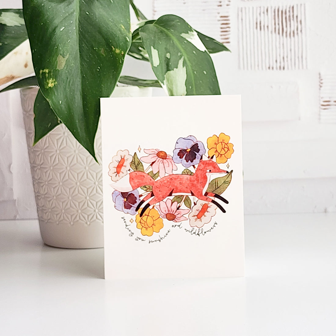 greeting card with an illustrated fox jumping through blooming flowers and the words sending you sunshine and wildflowers. The card is on a white background with a potted plant behind it