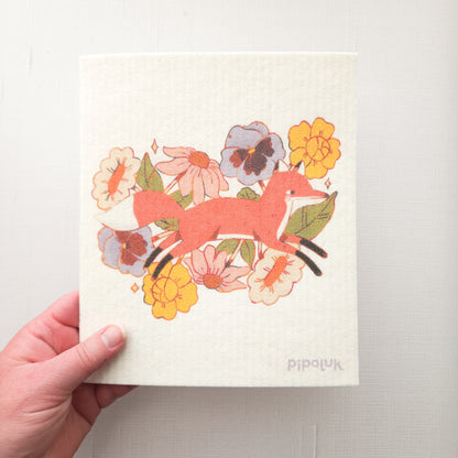 swedish dishcloth with an image of a fox jumping through flowers and a pipaluk logo in the bottom right corner, held in a hand