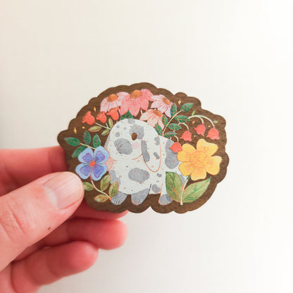 hand holding a wooden magnet with an illustration of a gray and white bunny surrounded by flowers on a linen background