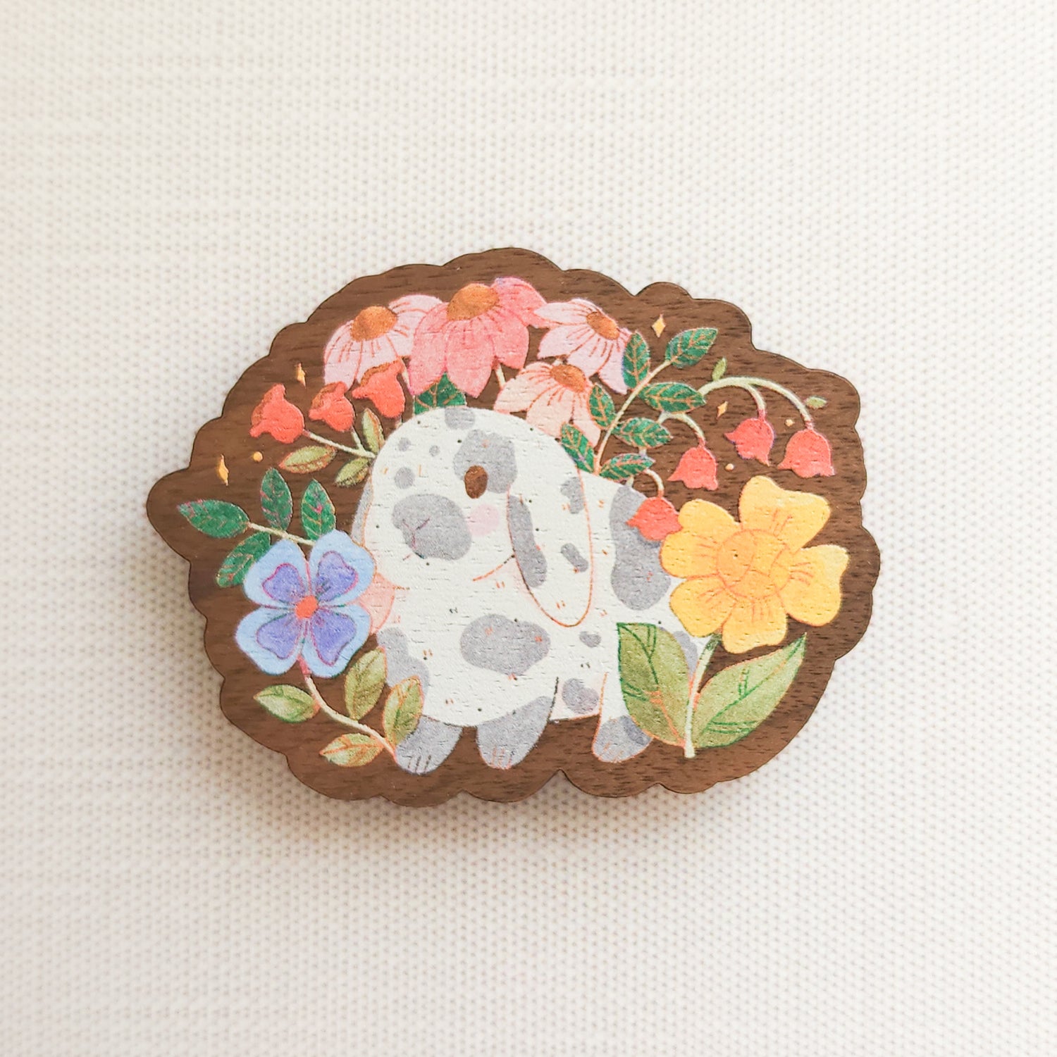 wooden magnet with an illustration of a gray and white bunny surrounded by flowers on a linen background