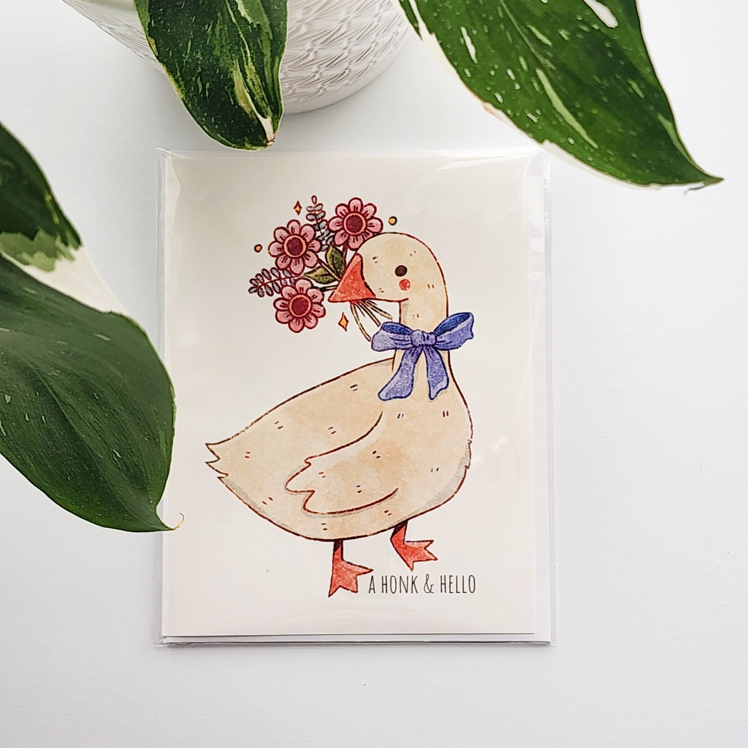 greeting card with an illustrated goose holding a bouquet of flowers. The card  sitting on a white background with a potted plant