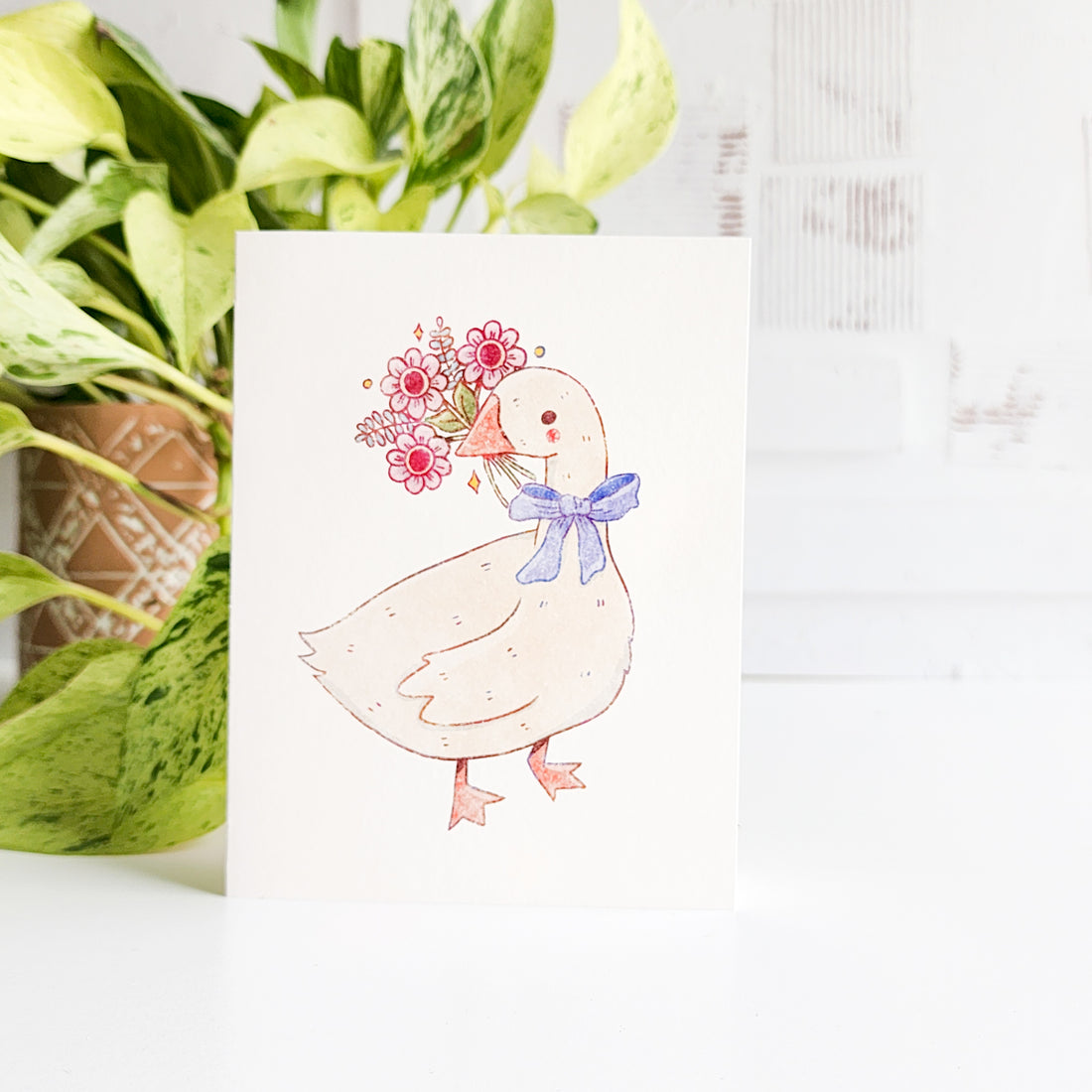 Card with a goose illustration holding flowers, placed next to a potted plant.
