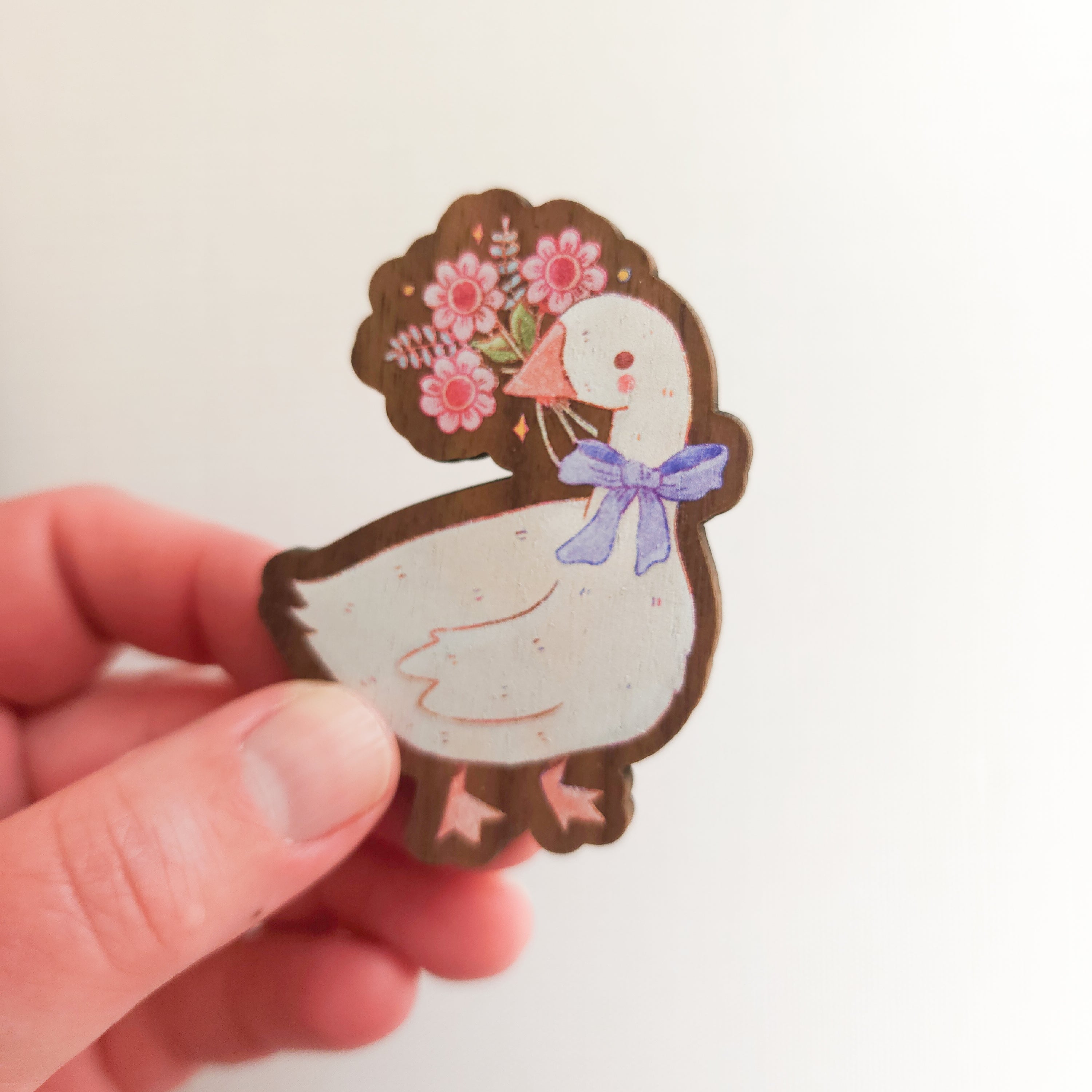 hand holding a wooden magnet with an illustration of a goose holding a bouquet of flowers in its beak on a linen background