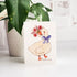greeting card with an illustrated goose holding a bouquet of flowers. The card  sitting on a white background with a potted plant