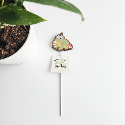 Small decorative plant stake with a frog design next to a potted plant on a white surface.
