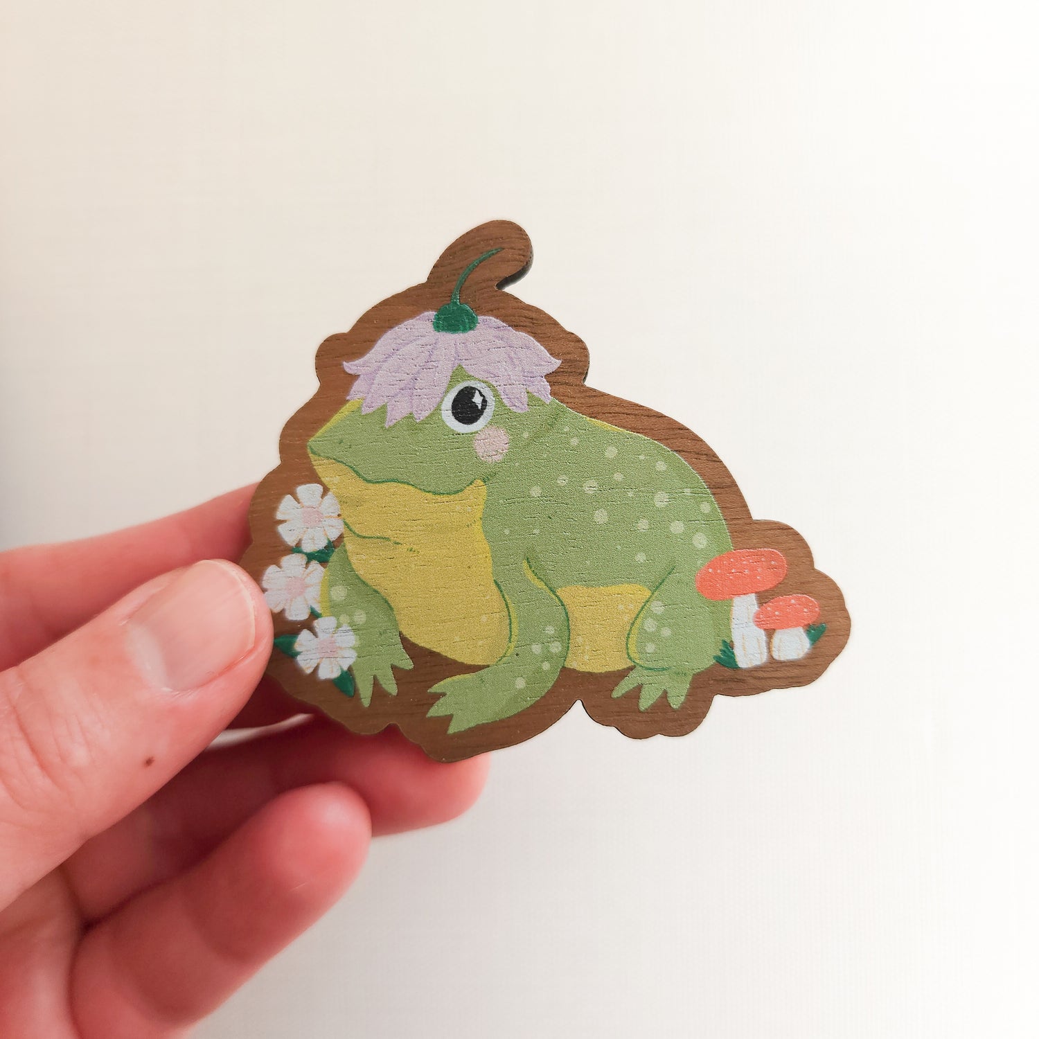 hand holding a wooden magnet with an illustration of a happy toad wearing a flower a hat on a linen background