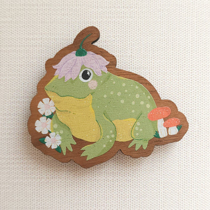 wooden magnet with an illustration of a happy toad wearing a flower a hat on a linen background