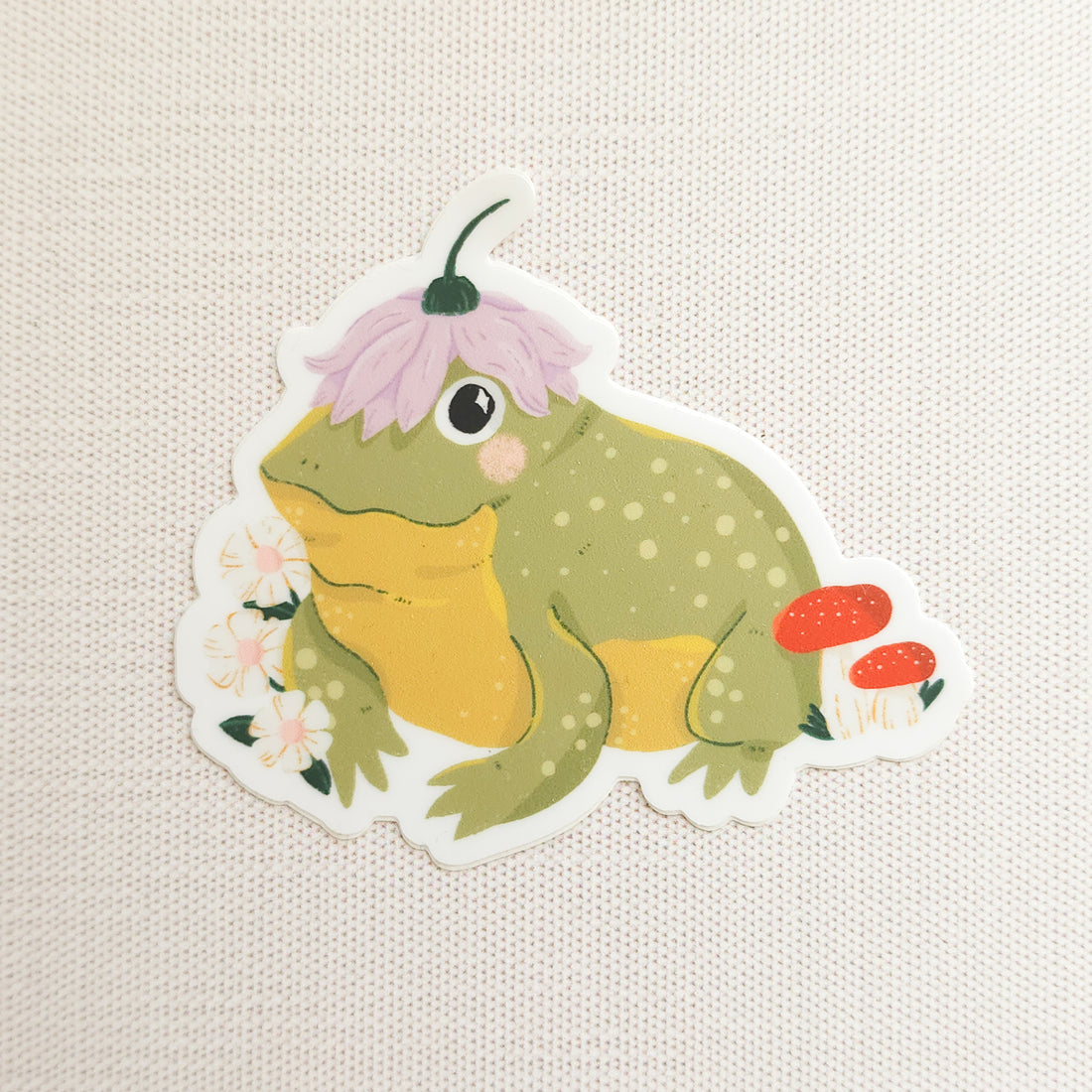 Illustrated sticker of a frog with a flower on its head on a linen background
