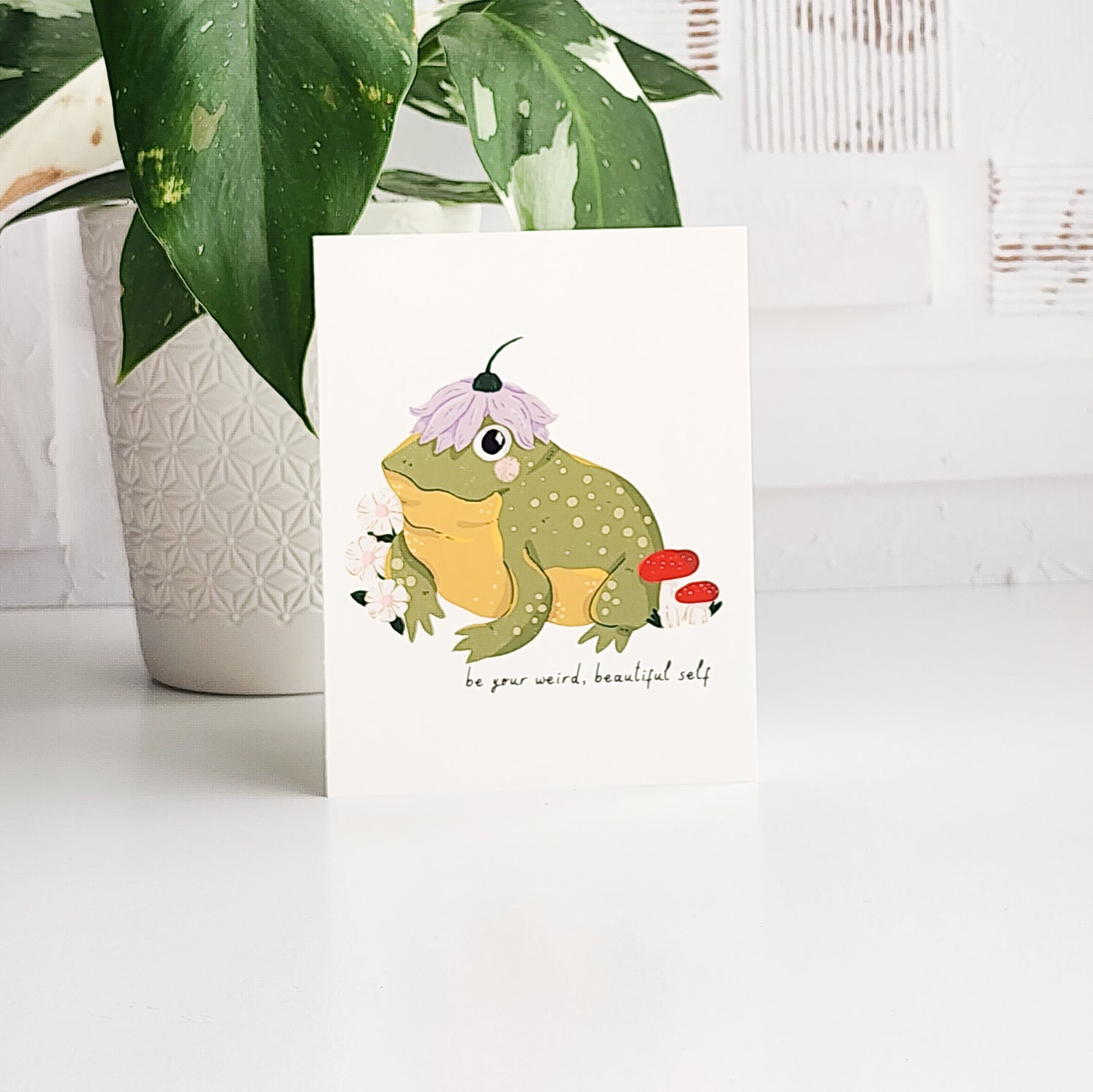 illustrated card of a toad wearing a flower hat and the words be your weird beautiful self. The card is on a white background with a potted plant