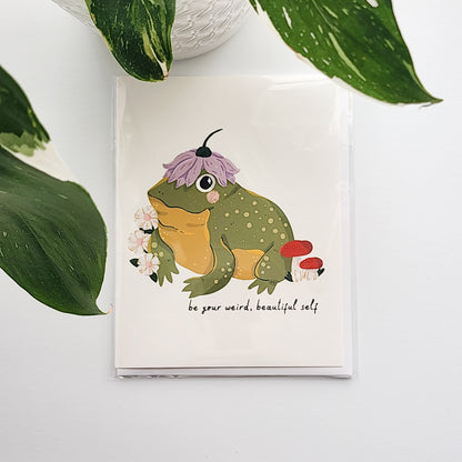 illustrated card of a toad wearing a flower hat and the words be your weird beautiful self. The card is on a white background with a potted plant