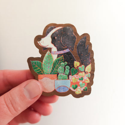 hand holding a wooden magnet with an illustration of a black and white dog sitting behind potted houseplants on a linen background