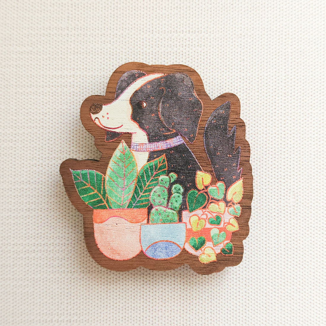 wooden magnet with an illustration of a black and white dog sitting behind potted houseplants on a linen background