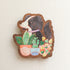 wooden magnet with an illustration of a black and white dog sitting behind potted houseplants on a linen background
