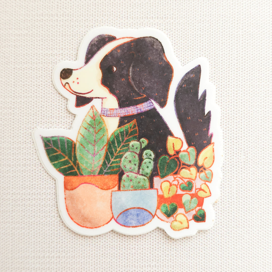 sticker with an illustration of a black and white dog sitting behind potted plants on a linen background