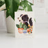 Card with a dog illustration surrounded by plants on a white surface with a potted plant