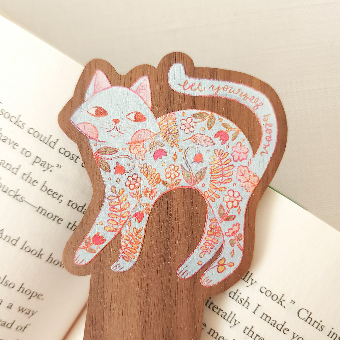 wooden bookmark with a floral cat illustration and the words let yourself bloom sitting on an open book
