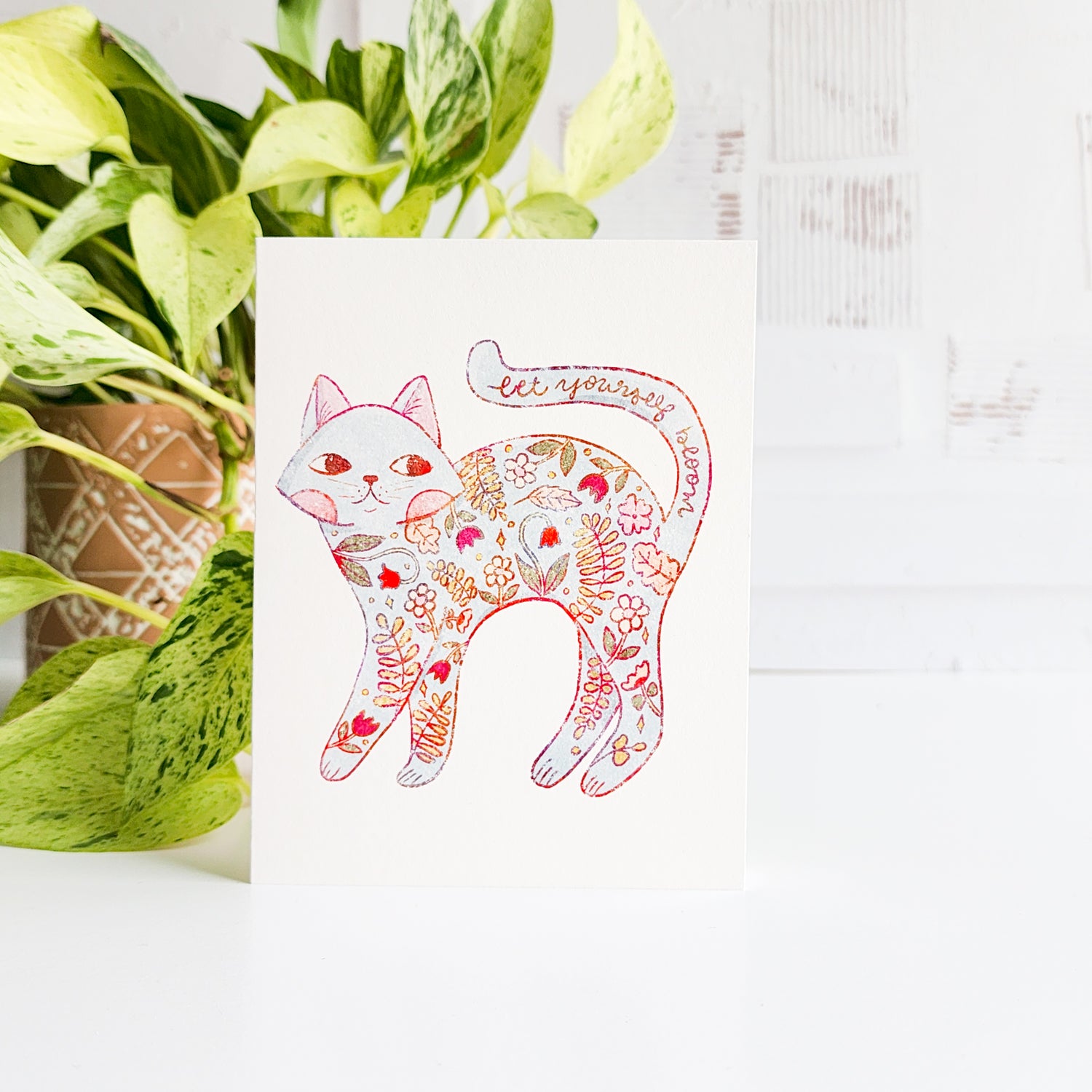 Decorative card with a floral cat design next to a potted plant on a white surface.