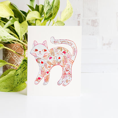 Decorative card with a floral cat design next to a potted plant on a white surface.