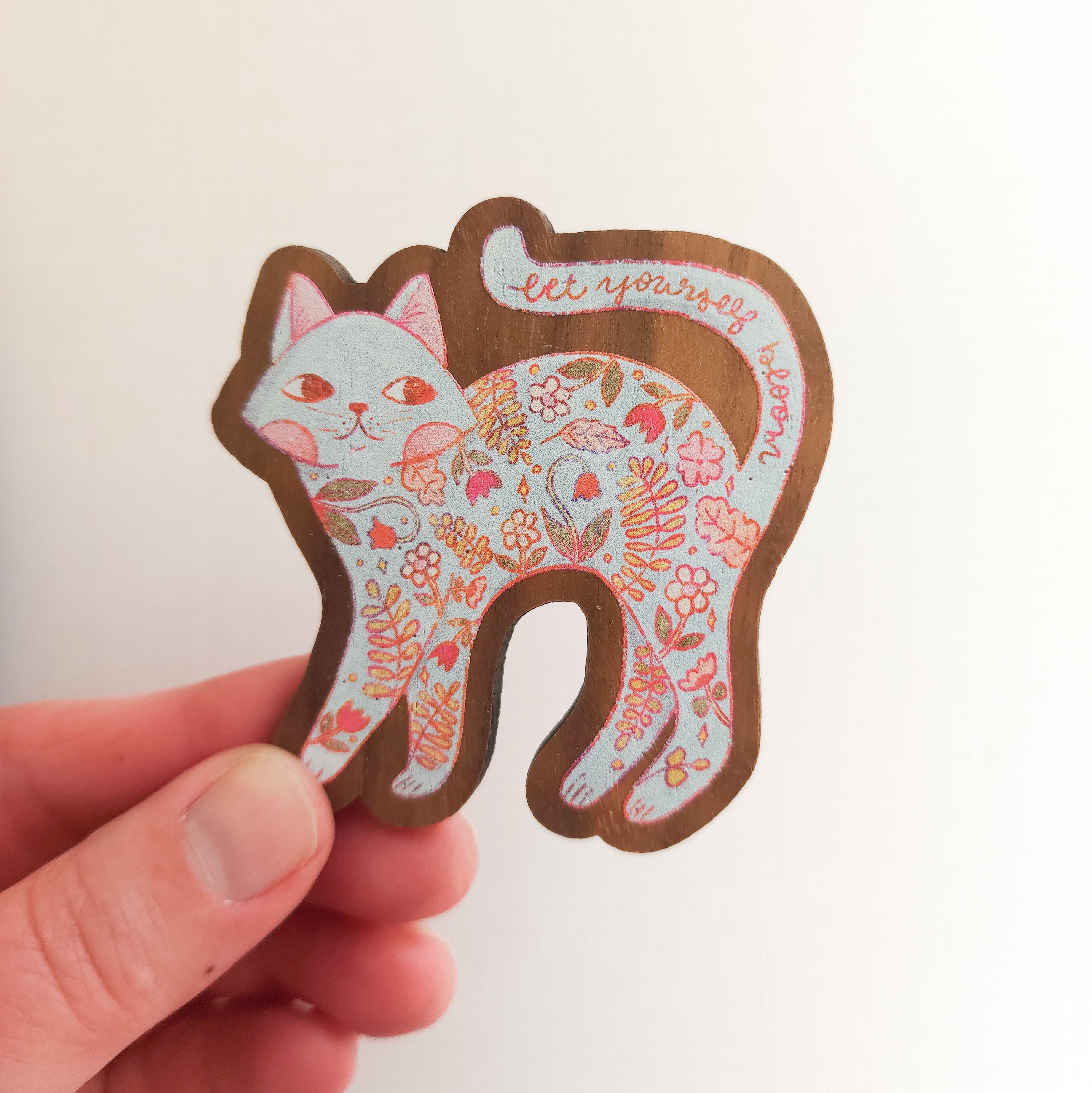 hand holding a wooden magnet with an illustration a a cat with flowers inside and the words let yourself bloom on a linen background