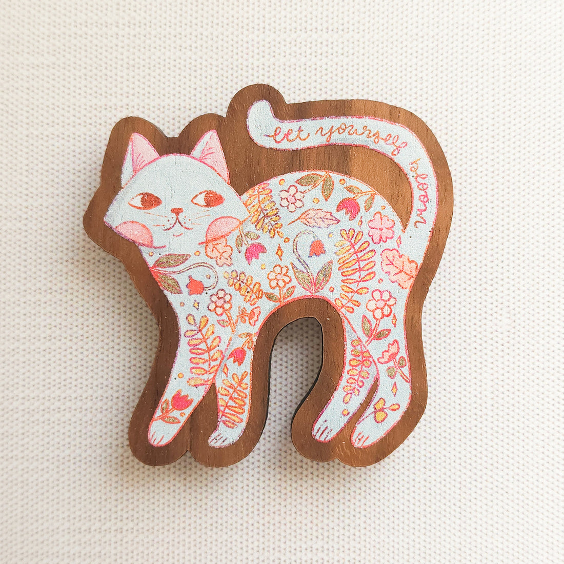 wooden magnet with an illustration a a cat with flowers inside and the words let yourself bloom on a linen background