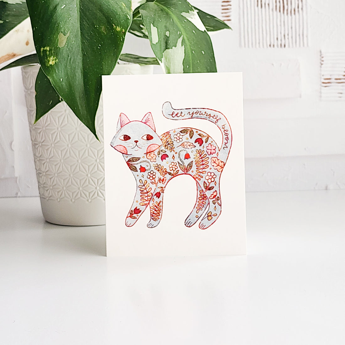 card with an illustrated floral cat and the words let yourself bloom on a white background in front of a plant 