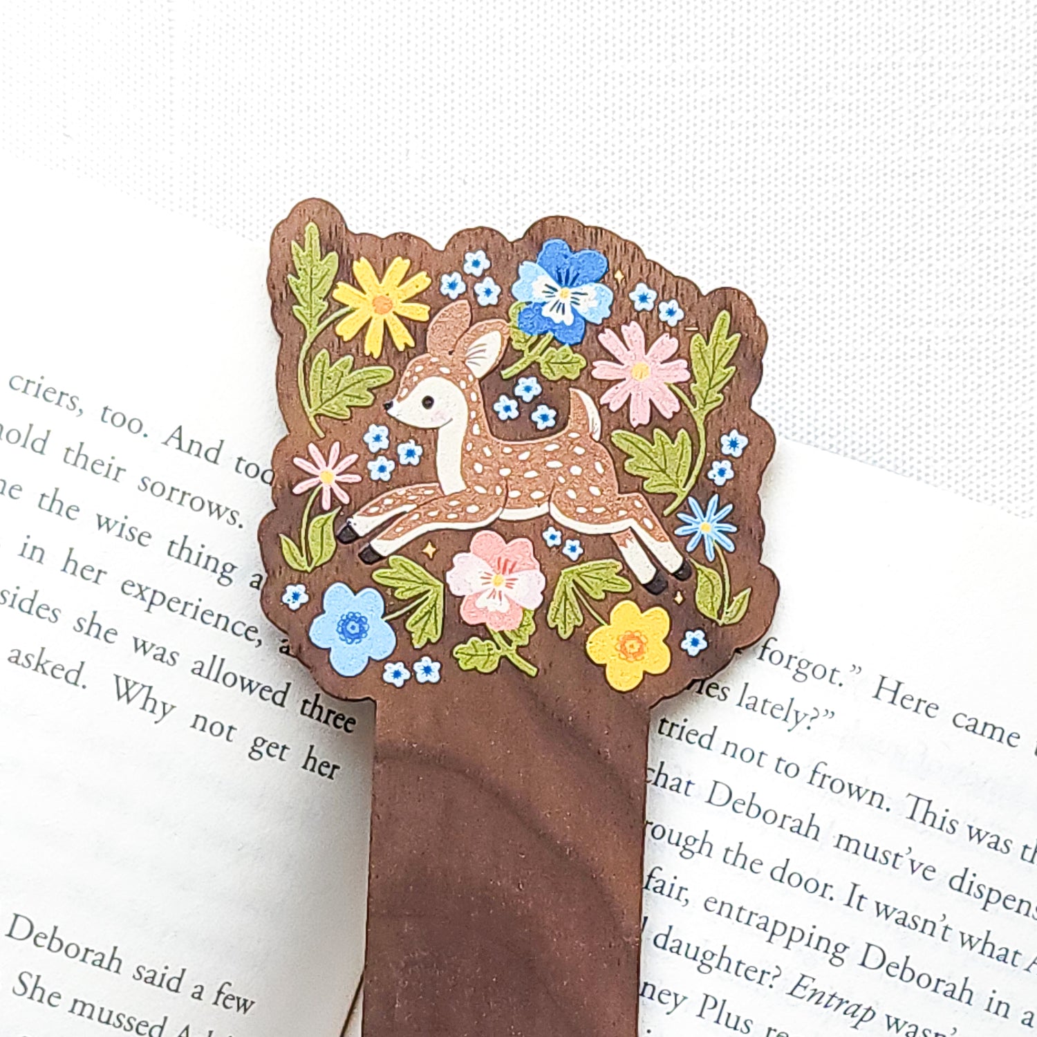 Wooden bookmark featuring a sweet spotted fawn dancing among colorful wildflowers, printed on walnut wood and photographed on top of a book on a neutral fabric background