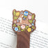 Wooden bookmark featuring a sweet spotted fawn dancing among colorful wildflowers, printed on walnut wood and photographed on top of a book on a neutral fabric background
