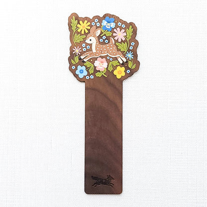 Wooden bookmark featuring a sweet spotted fawn dancing among colorful wildflowers, printed on walnut wood and photographed on a neutral fabric background.