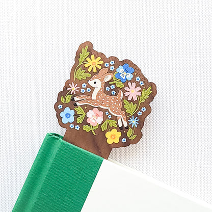 Wooden bookmark featuring a sweet spotted fawn dancing among colorful wildflowers, printed on walnut wood and photographed in a book on a neutral fabric background.