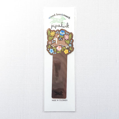 Wooden bookmark featuring a sweet spotted fawn dancing among colorful wildflowers, printed on walnut wood and photographed in Pipaluk packaging on a neutral fabric background