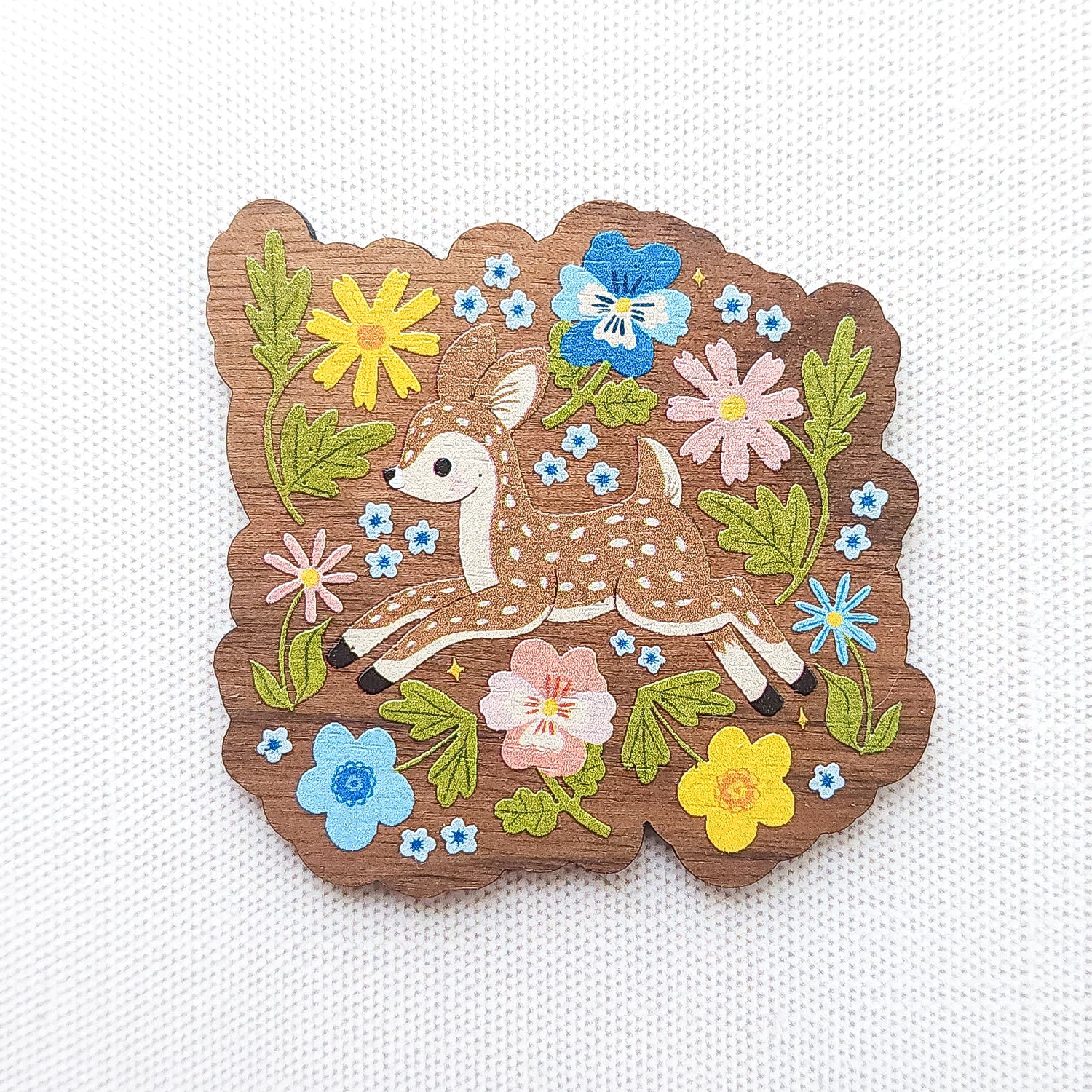 Wooden magnet featuring a sweet spotted fawn dancing among colorful wildflowers, printed on walnut wood and photographed on a neutral fabric background.