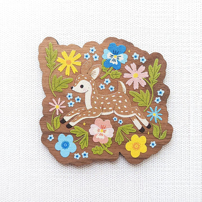 Wooden magnet featuring a sweet spotted fawn dancing among colorful wildflowers, printed on walnut wood and photographed on a neutral fabric background.