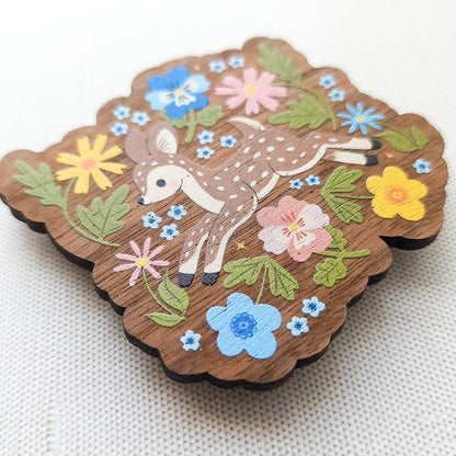 Wooden magnet featuring a sweet spotted fawn dancing among colorful wildflowers, printed on walnut wood and photographed on a neutral fabric background.