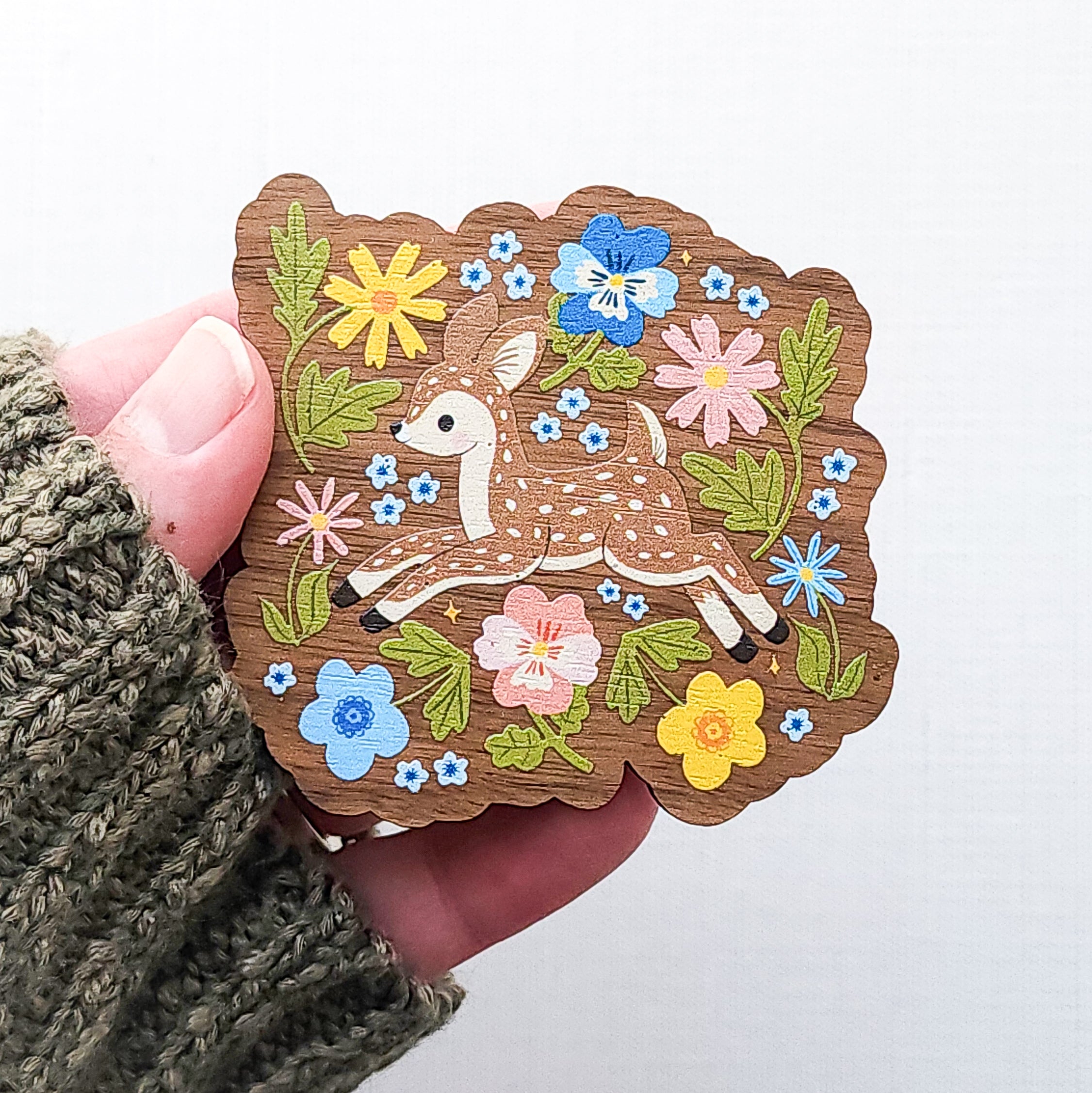 Wooden magnet featuring a sweet spotted fawn dancing among colorful wildflowers, printed on walnut wood and photographed held in a hand on a neutral fabric background.