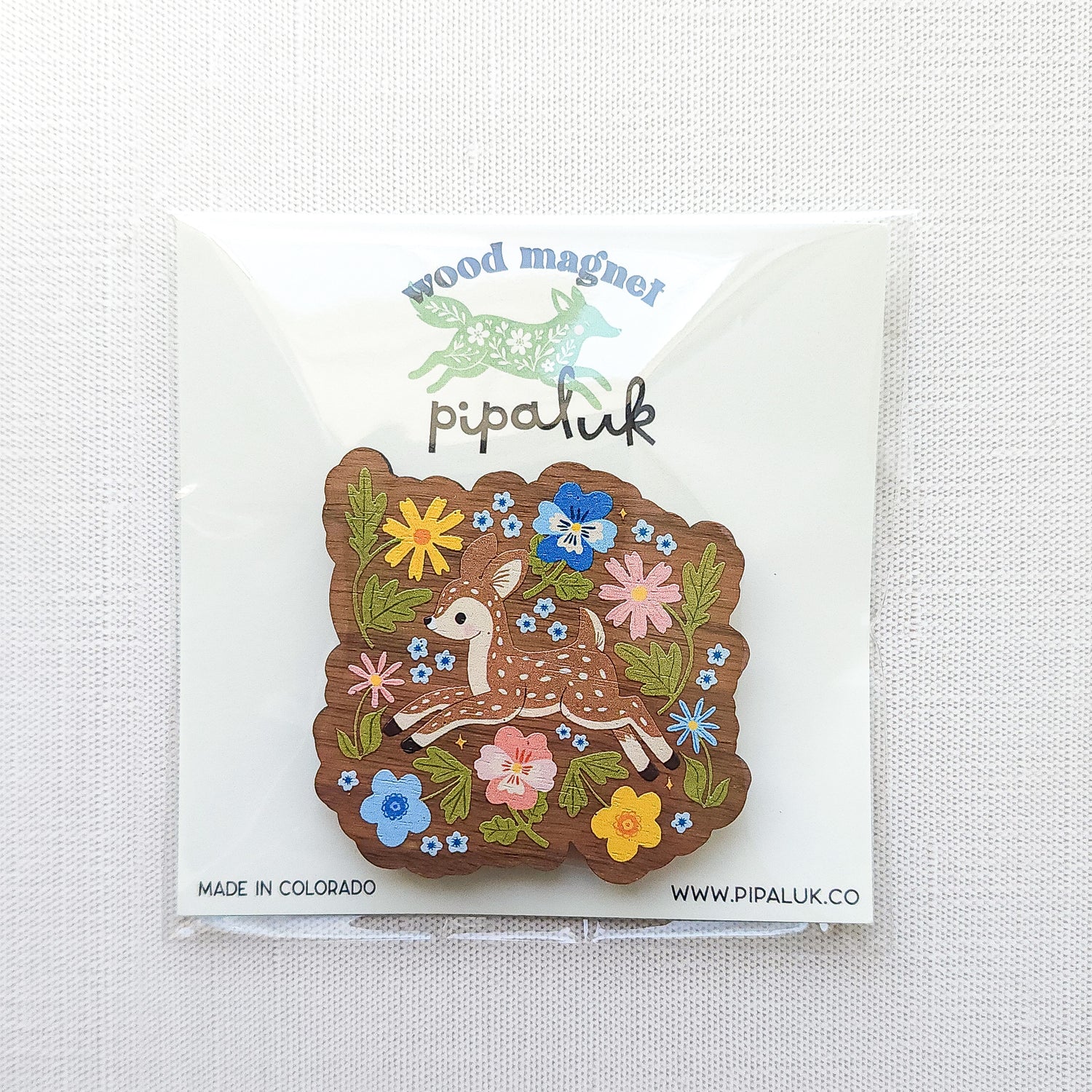 Wooden magnet featuring a sweet spotted fawn dancing among colorful wildflowers, printed on walnut wood and photographed in Pipaluk packaging on  a neutral fabric background.