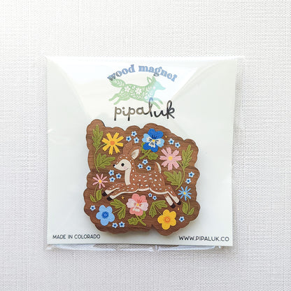 Wooden magnet featuring a sweet spotted fawn dancing among colorful wildflowers, printed on walnut wood and photographed in Pipaluk packaging on  a neutral fabric background.