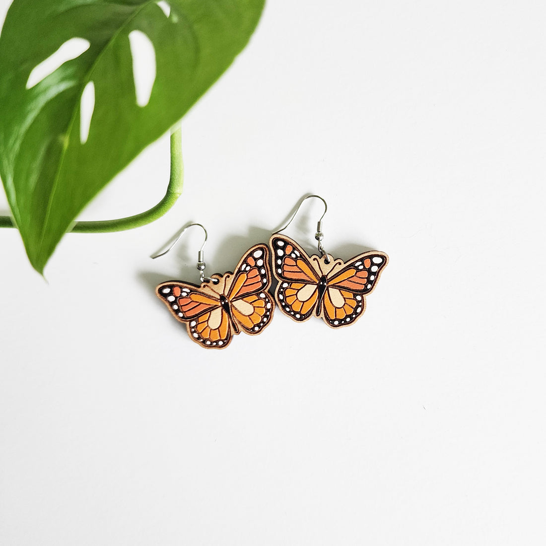 pair of monarch butterfly earrings on a white background next to a leaf