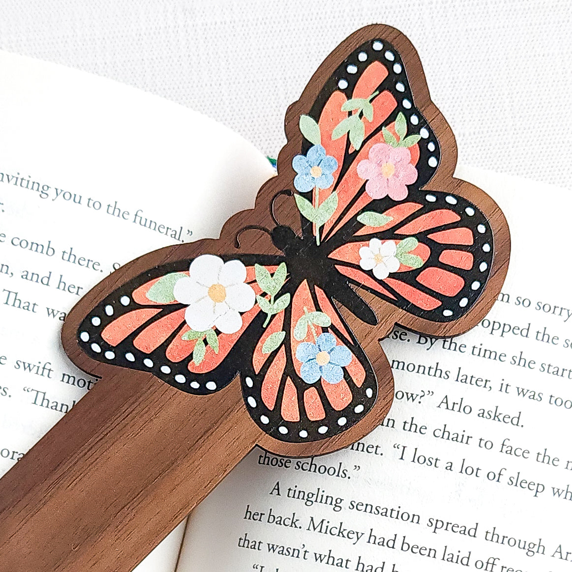 Wooden bookmark featuring a monarch butterfly with orange wings, black detailing, and tiny colorful blooms, printed on walnut wood and photographed on an open book and neutral fabric background.