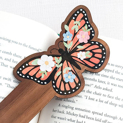 Wooden bookmark featuring a monarch butterfly with orange wings, black detailing, and tiny colorful blooms, printed on walnut wood and photographed on an open book and neutral fabric background.