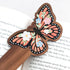 Wooden bookmark featuring a monarch butterfly with orange wings, black detailing, and tiny colorful blooms, printed on walnut wood and photographed on an open book and neutral fabric background.