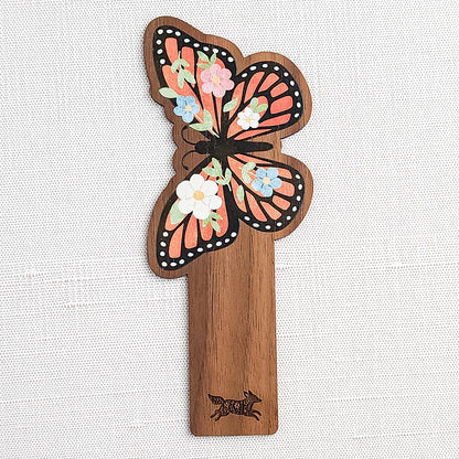 Wooden bookmark featuring a monarch butterfly with orange wings, black detailing, and tiny colorful blooms, printed on walnut wood and photographed on a neutral fabric background.