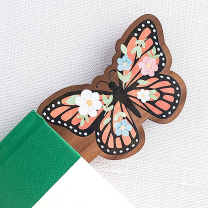 Wooden bookmark featuring a monarch butterfly with orange wings, black detailing, and tiny colorful blooms, printed on walnut wood and photographed inside of a book on a neutral fabric background.