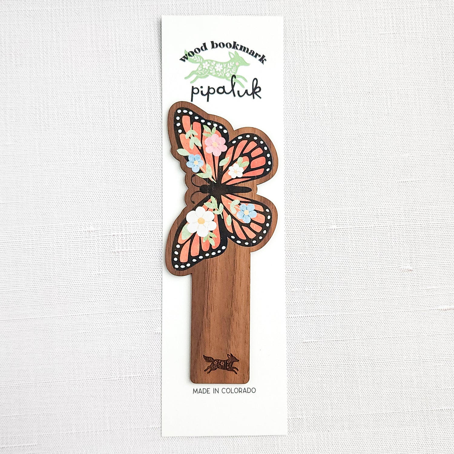 Wooden bookmark featuring a monarch butterfly with orange wings, black detailing, and tiny colorful blooms, printed on walnut wood and photographed in Pipaluk branded packaging  on a neutral fabric background.