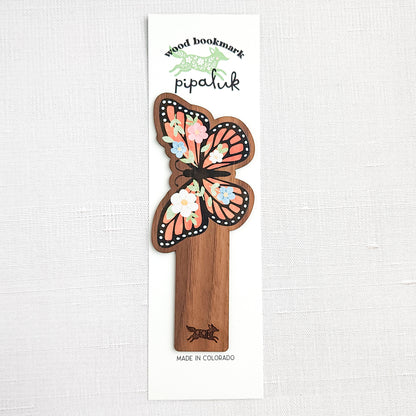 Wooden bookmark featuring a monarch butterfly with orange wings, black detailing, and tiny colorful blooms, printed on walnut wood and photographed in Pipaluk branded packaging  on a neutral fabric background.