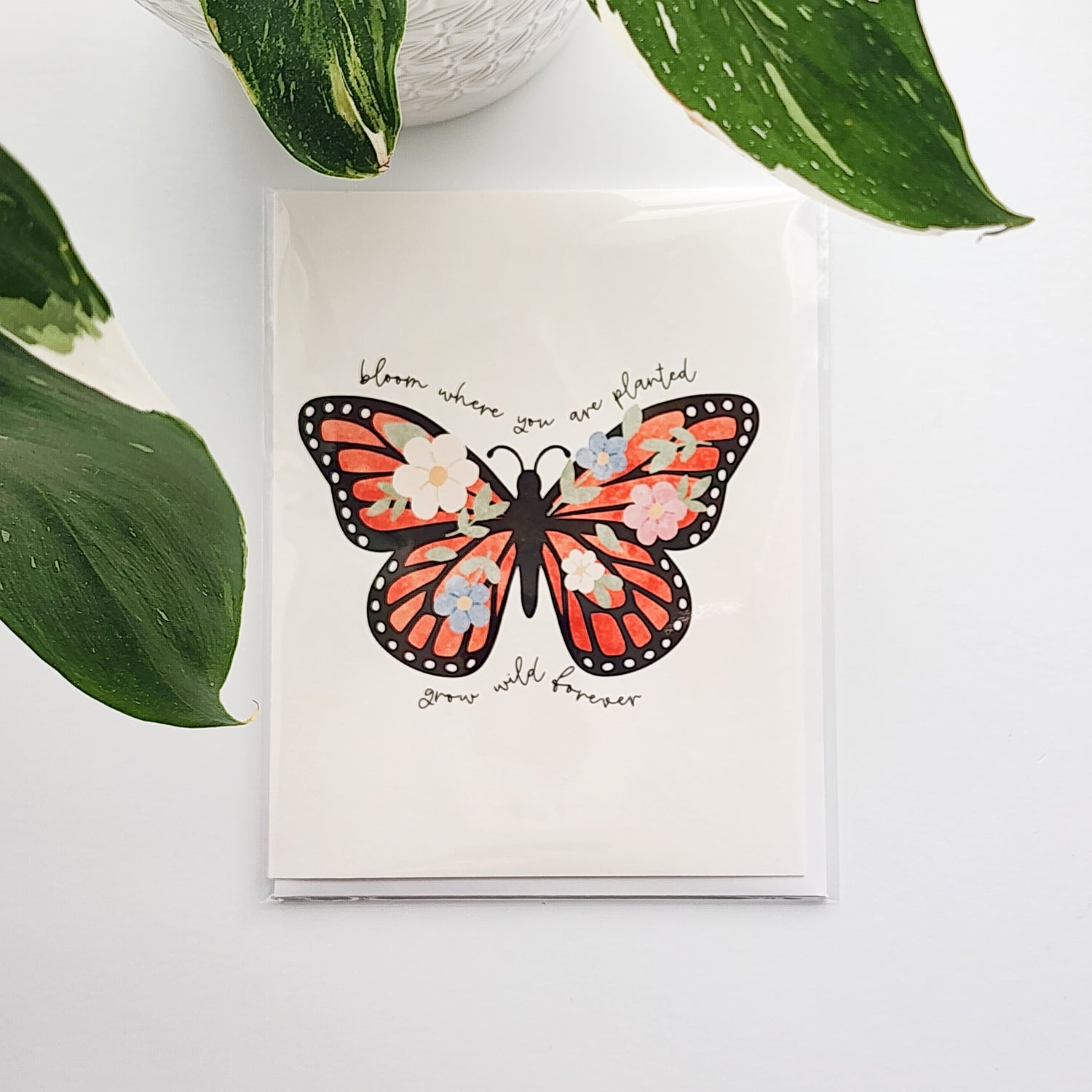 greeting card with an illustrated butterfly and the words bloom where you are planted, grow wild forever. The card is on a white background next to a potted plant